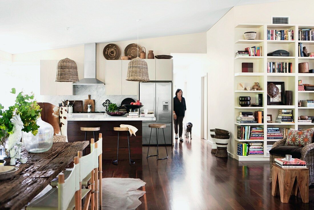 Moderner, offener Wohnraum mit Regalwand, Essplatz mit rustikaler Holzplatte, im Hintergrund Küchenbereich mit Korblampen und Frau mit Hund