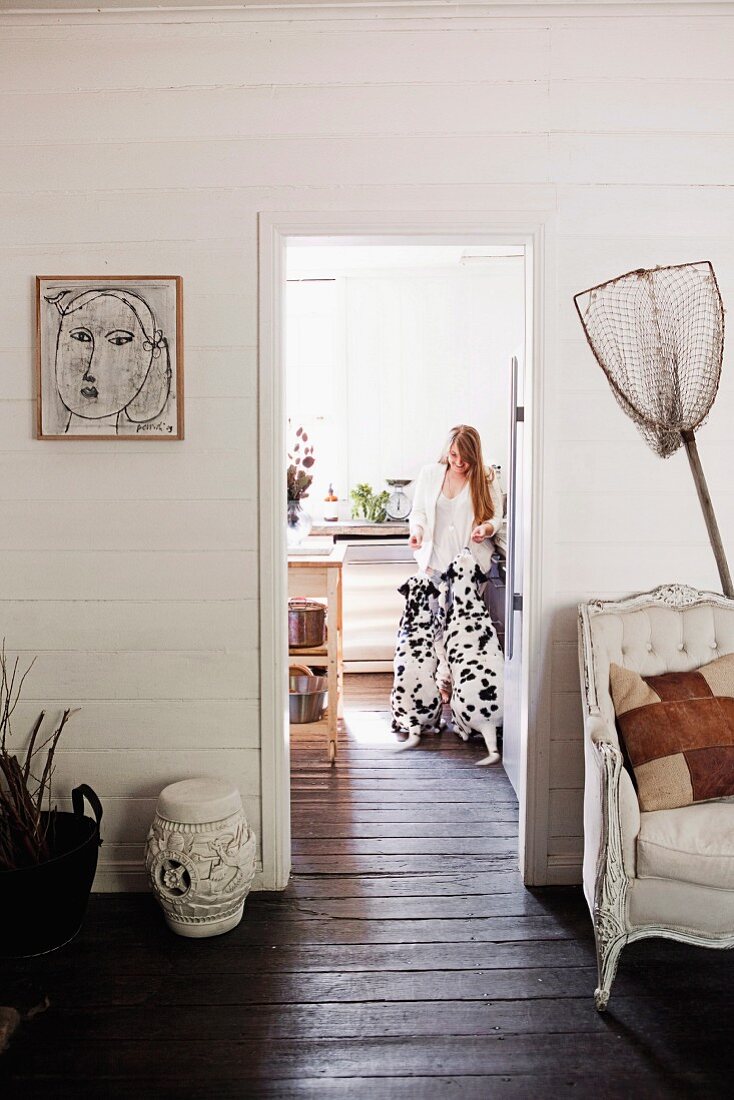 Blick vom Vorraum mit Fischernetz und Vintage Sessel an weisser Holzwand, in Nebenraum mit durchgehendem, dunkelbraunem Dielenboden, Frau mit zwei Dalmatiner in Küche
