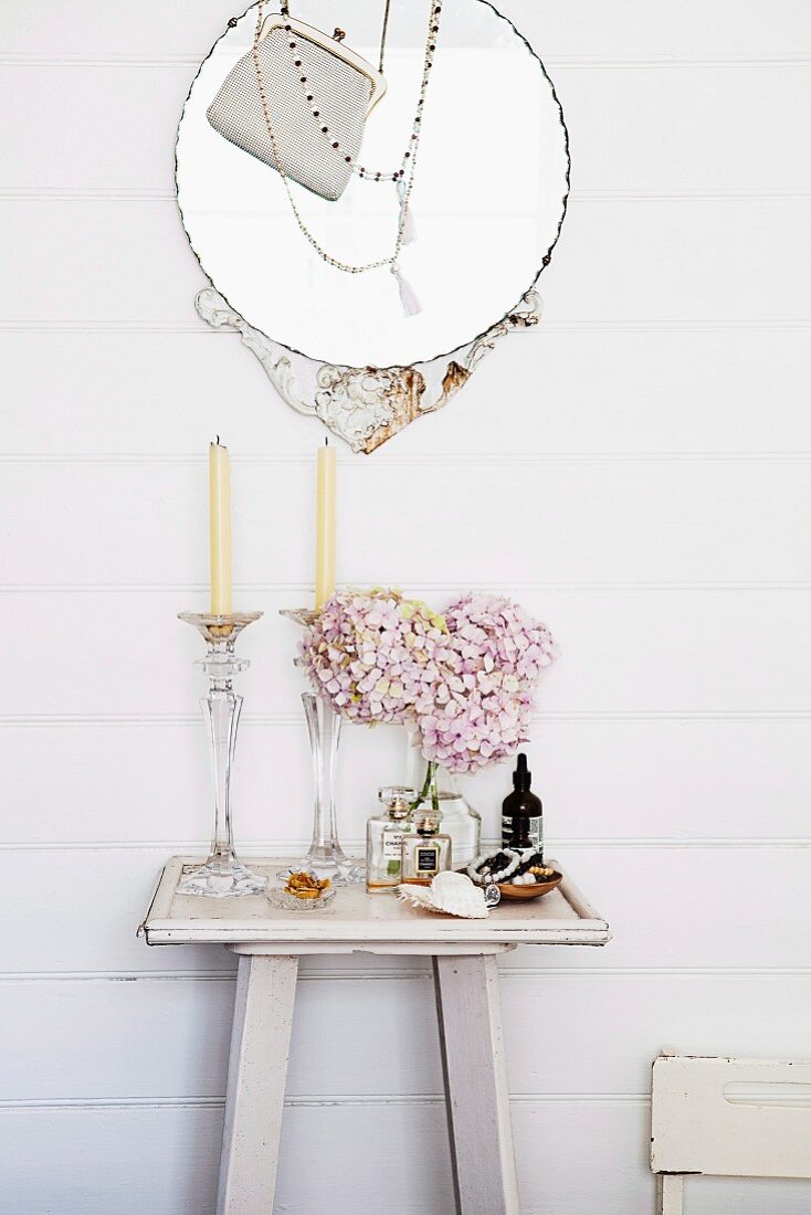 Glas Kerzenständer und rosa Hortensienblüten in Vase, auf weisslackierter Wandkonsole, darüber mit Halskette und Abendtäschchen dekorierter, runder Spiegel an Holzwand