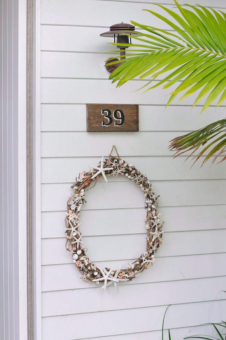 Delicate wreath below house number and lamp on white wooden façade behind palm frond