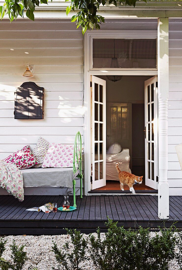 Weisses Holzhaus mit offener Terrassentür, Katze spaziert hinaus; gemütliches Vintage Tagesbett mit geblümten Kissen auf schwarzem Holzboden