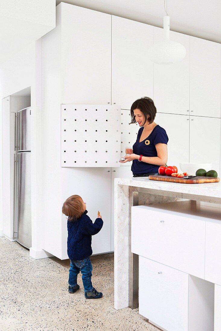 Mutter mit Kind in offener Küche, Küchentheke aus Stein und eingeschobenen, weissen Schrankmodulen; seitlich raumhoher, weisser Einbauschrank