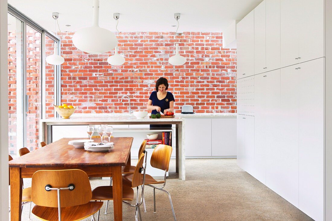 Essbereich vor Küchentheke mit Frau bei Essensvorbereitung, im Hintergrund Ziegelwand; seitlich hoher weißer Einbauschrank in moderner offener Küche mit Klassiker Stühlen