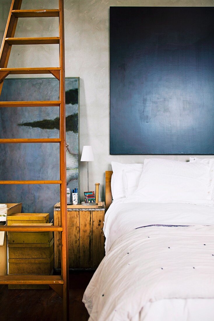 Bed with white bed linen below dark painting next to long wooden ladder and wooden box used as bedside table