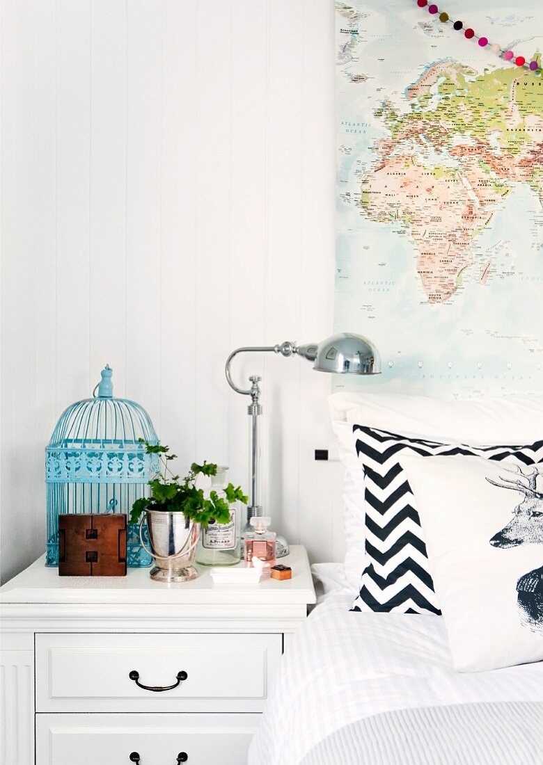 Weißes Schubladennachtkästchen dekoriert mit hellblauem Vogelkäfig und nostalgischer Nachttischlampe, Bett mit Zickzackmuster-Kissen und aufgehängter Landkarte