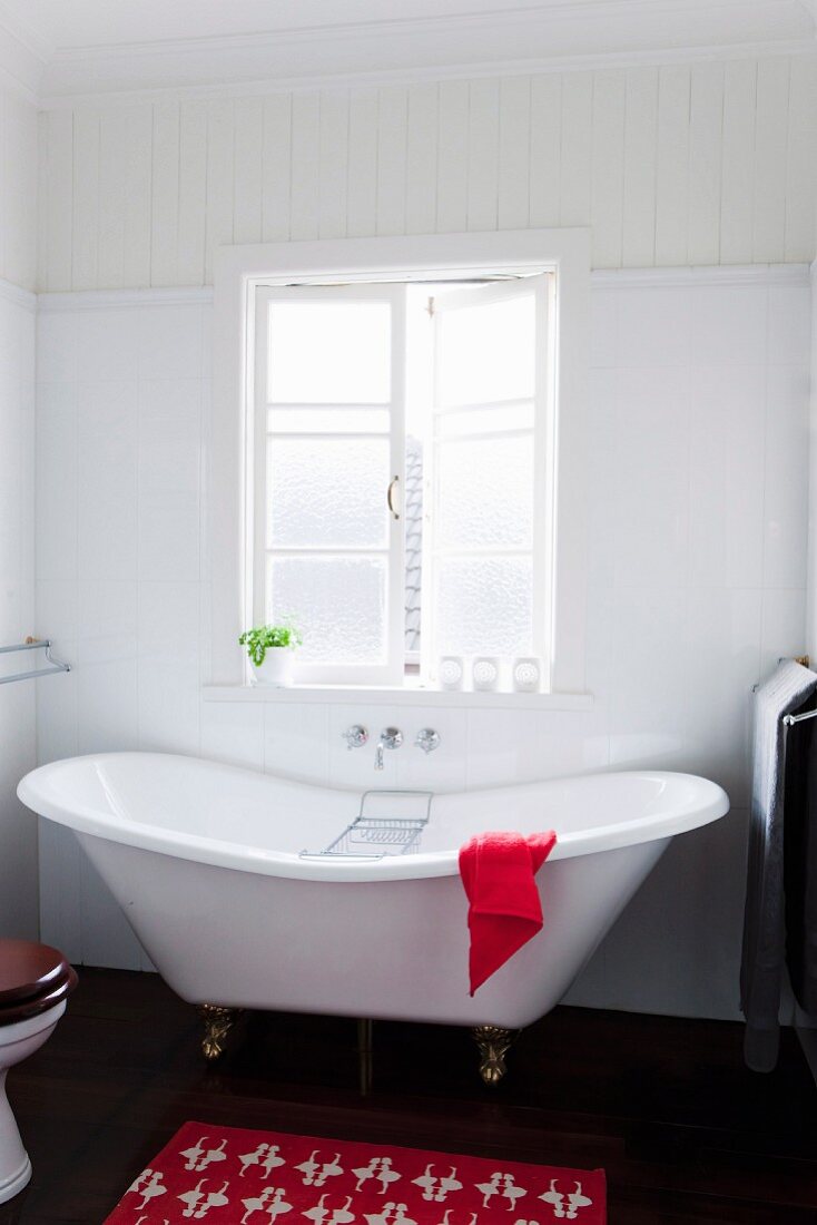 Red bathmat on dark wooden floor beside vintage-style white bathtub below lattice window