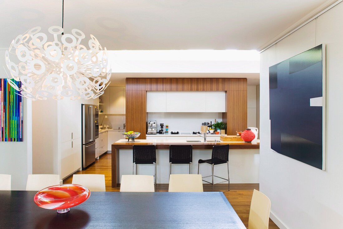 Designer Dandelion pendant lamp, colourful 60s glass bowl and dark dining table in front of breakfast bar in open-plan kitchen
