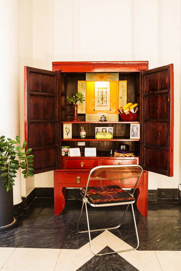 Klappstuhl vor rotem chinesischem Hochzeitsschrank mit Blick durch weitgeöffnete Türen auf die Innendekoration in der Art eines Schreins