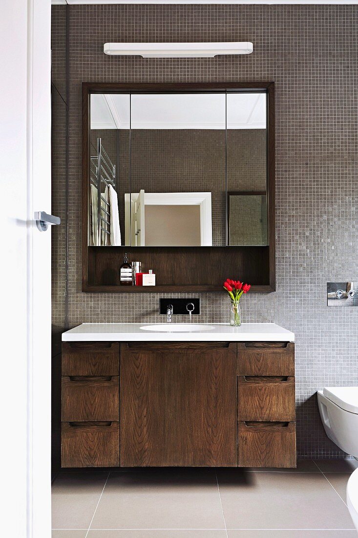 Washstand with wooden base unit and mirrored cabinet with three doors in bathroom with grey mosaic tiles