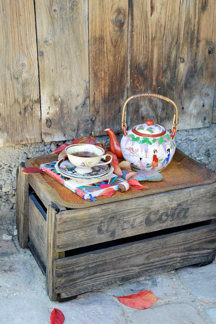 Rusty tray, vintage teacup and Chinese china teapot on weathered wooden crate used as side table against rustic wooden wall