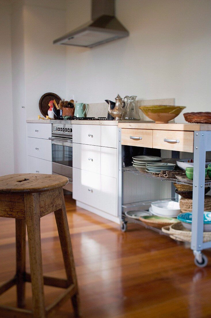 Rustic wooden stool, modern kitchen counter, crockery on trolley with drawers, white drawer units and stainless steel cooker under extractor fan