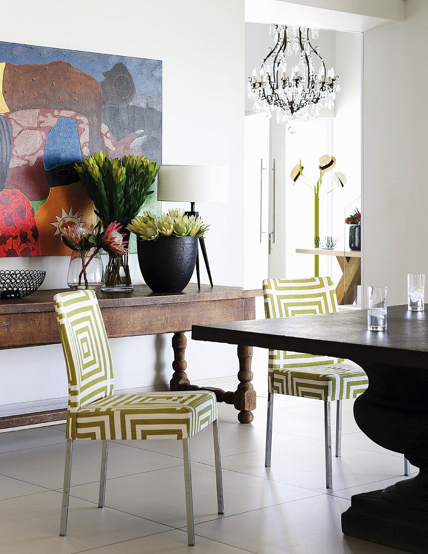 Stühle mit grafischem Muster auf weissgrünem Stoffbezug am Tisch, dahinter rustikaler Wandtisch mit Blumen in Vasen vor Bild an Wand, daneben raumhohe Öffnung und Blick auf Garderobenständer