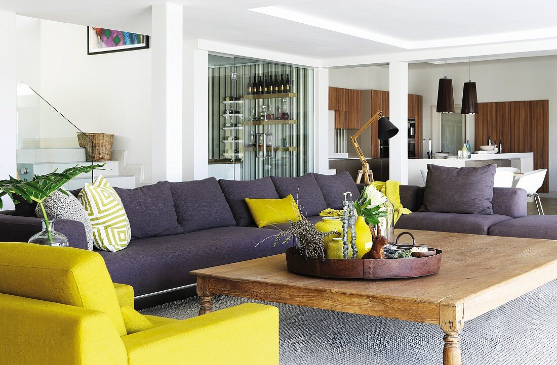 Grauviolettes Sofa und gelber Sessel um rustikalem Holz Couchtisch in grosszügigem Wohnraum mit Stützen, im Hintergrund Essbereich