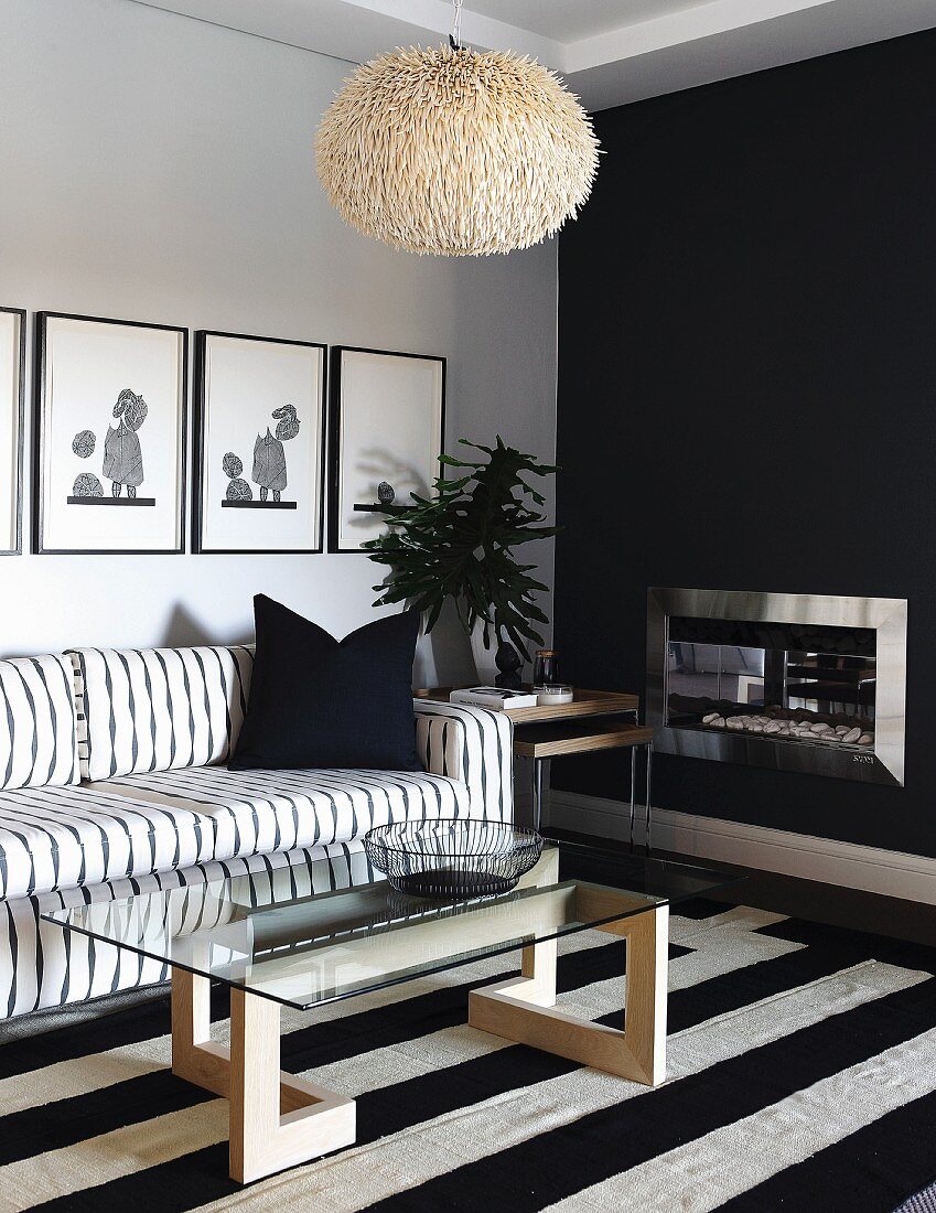 Coffee table with glass top on wooden base and black and white striped rug in front of black and white sofa below pictures on wall; open fireplace in black wall to one side