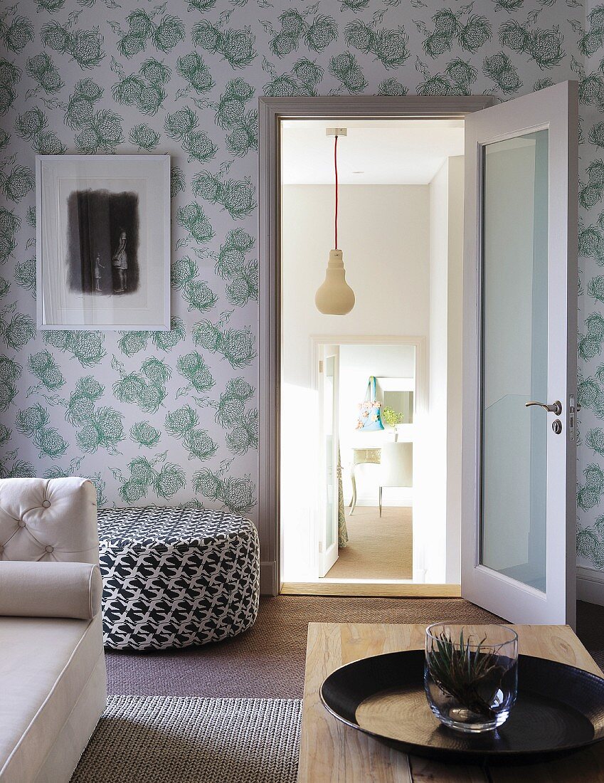 Wohnzimmer mit teilweise sichtbarem Couchtisch aus Holz und Sofa, dahinter schwarzweiss gemusterter Pouf an floral gemusterter Tapete an Wand, Blick durch offene Tür in tiefergelegte Nebenräume
