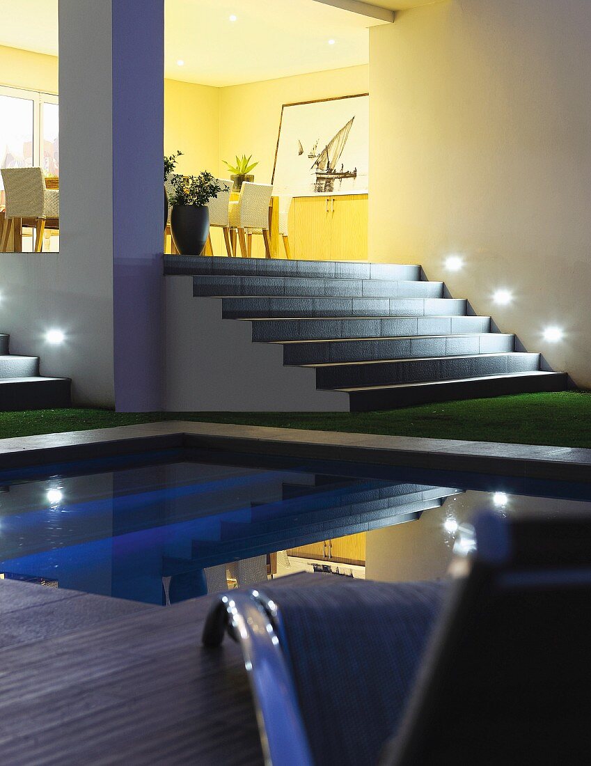 Pool vor breitem beleuchtetem Treppenaufgang, auf Podest Essplatz mit gepolsterten Stühlen in gelb getöntem Ambiente