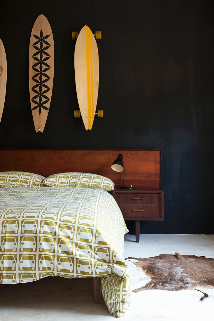 Doppelbett mit Kopfteil aus Holz und weissgrün gemusterte Bettwäsche mit Automotiven, an schwarzer Wand bemalte Skateboards aufgehängt