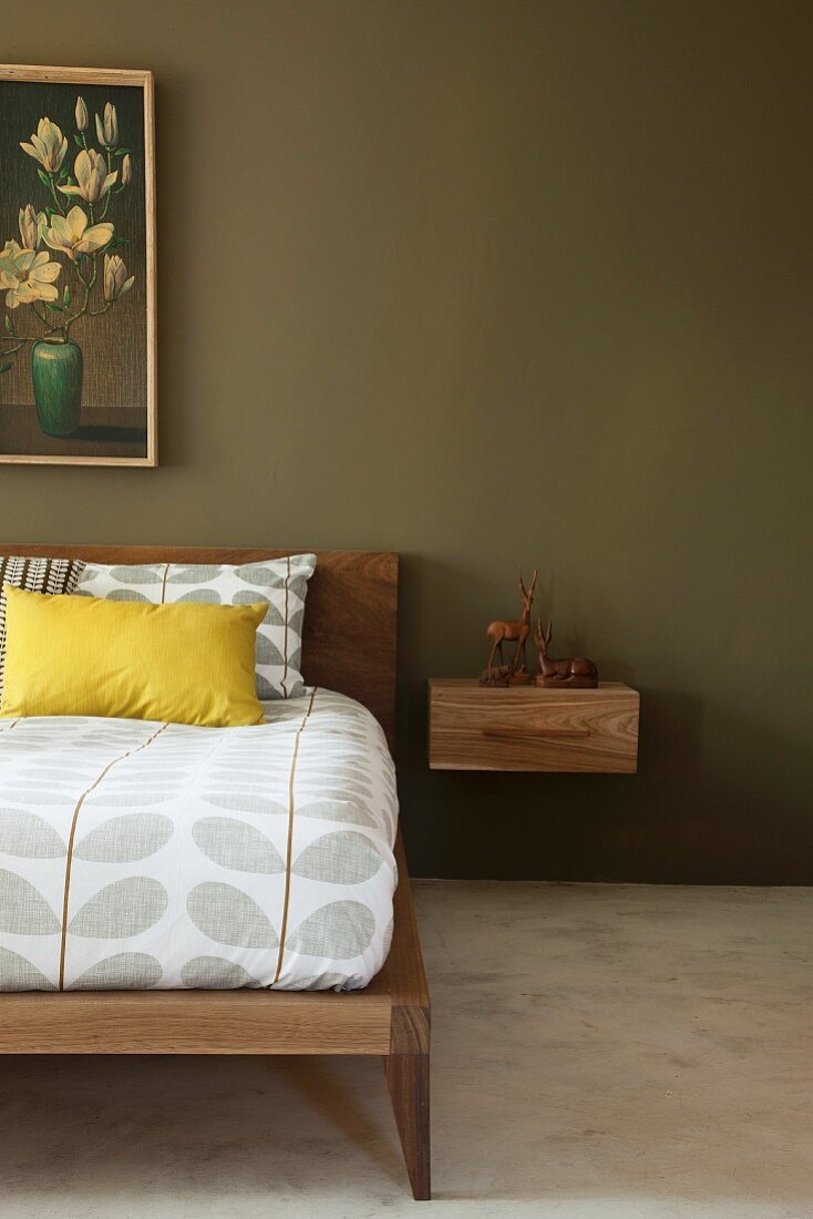 Bett mit Holzgestell und gemusterte Bettwäsche im Retrostil, neben gehängtem Nachtkästchen an grün getönter Wand mit teilweise sichtbarem Bild