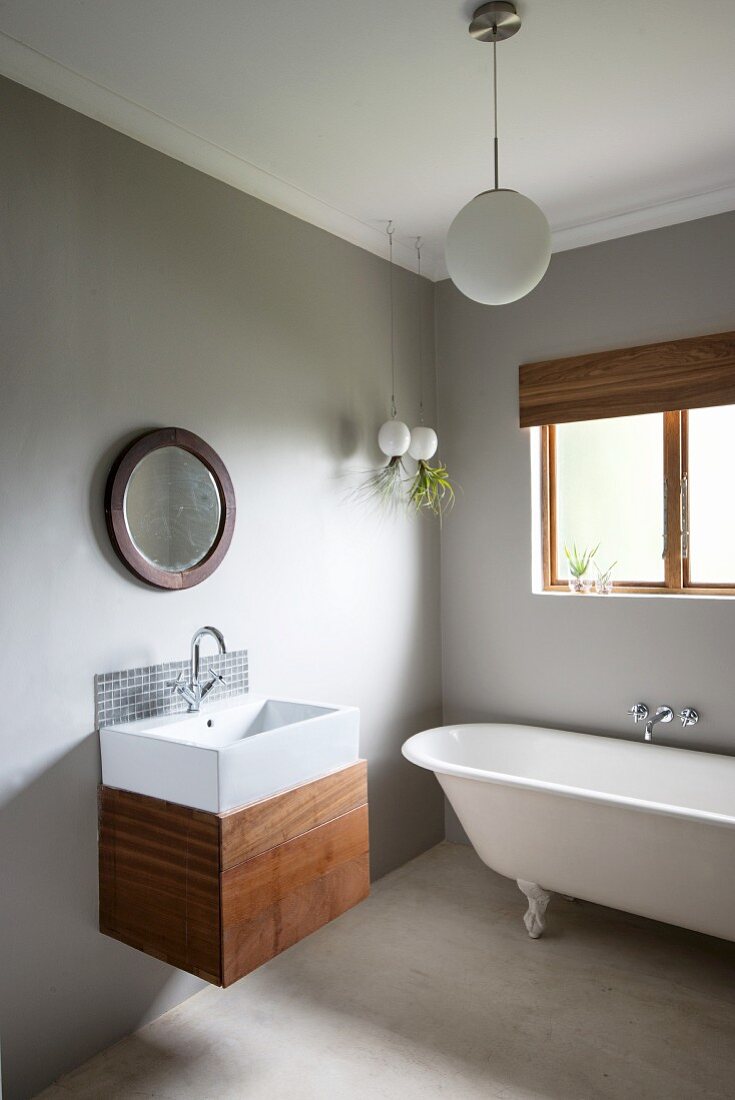 Modern sink with wooden base unit and minimalist, mosaic-tiled splashback below round mirror on wall painted pale grey; free-standing bathtub below window