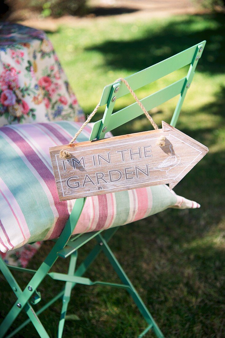 Gestreiftes Kissen auf pastellgrünen Gartenstuhl, Schild mit Info auf Rückenlehne gehängt