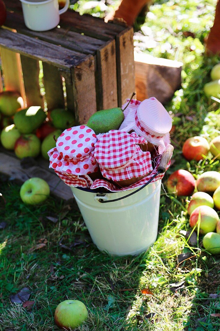 Vintage Holzkiste mit geernteten Äpfeln und Email-Eimer mit rot-weiß dekorierten Einmachgläsern im Garten