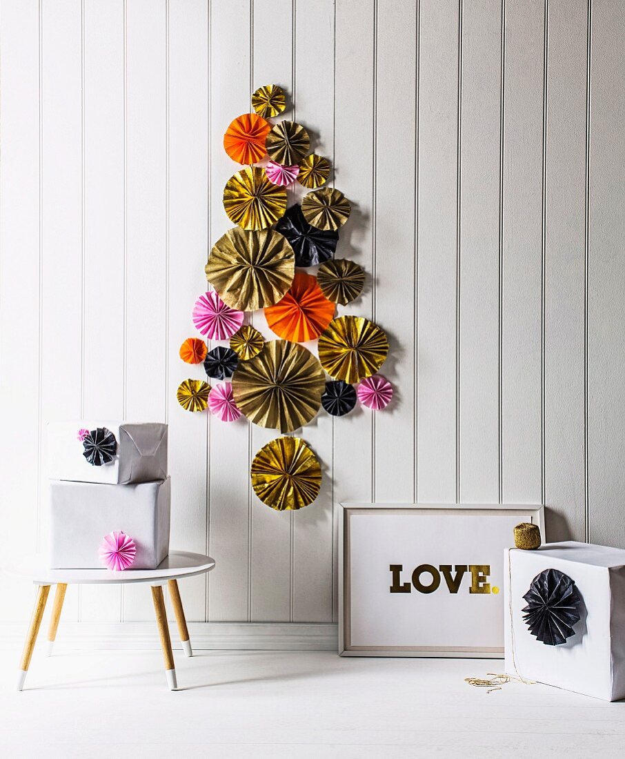 Verschiedene bunte Papierrossetten elegant als Weihnachtsbaum stilisiert an weißer Bretterwand befestigt, am Boden und Beistelltisch weiße Geschenkpakete arrangiert