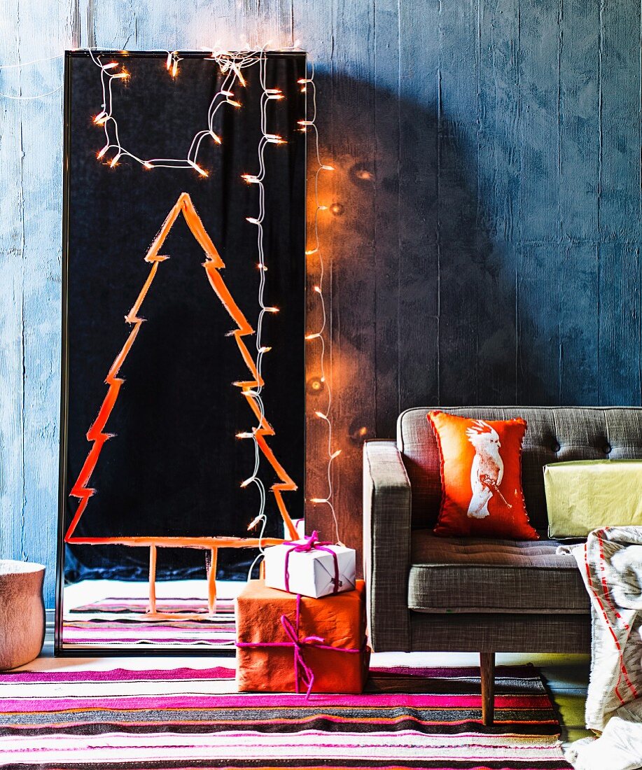 Bodenspiegel mit orangefarben gemalter Weihnachtsbaum Silhouette und Lichterkette an blaugrauer Wand, bunter Streifenteppich und Geschenkpakete neben Retrosofa