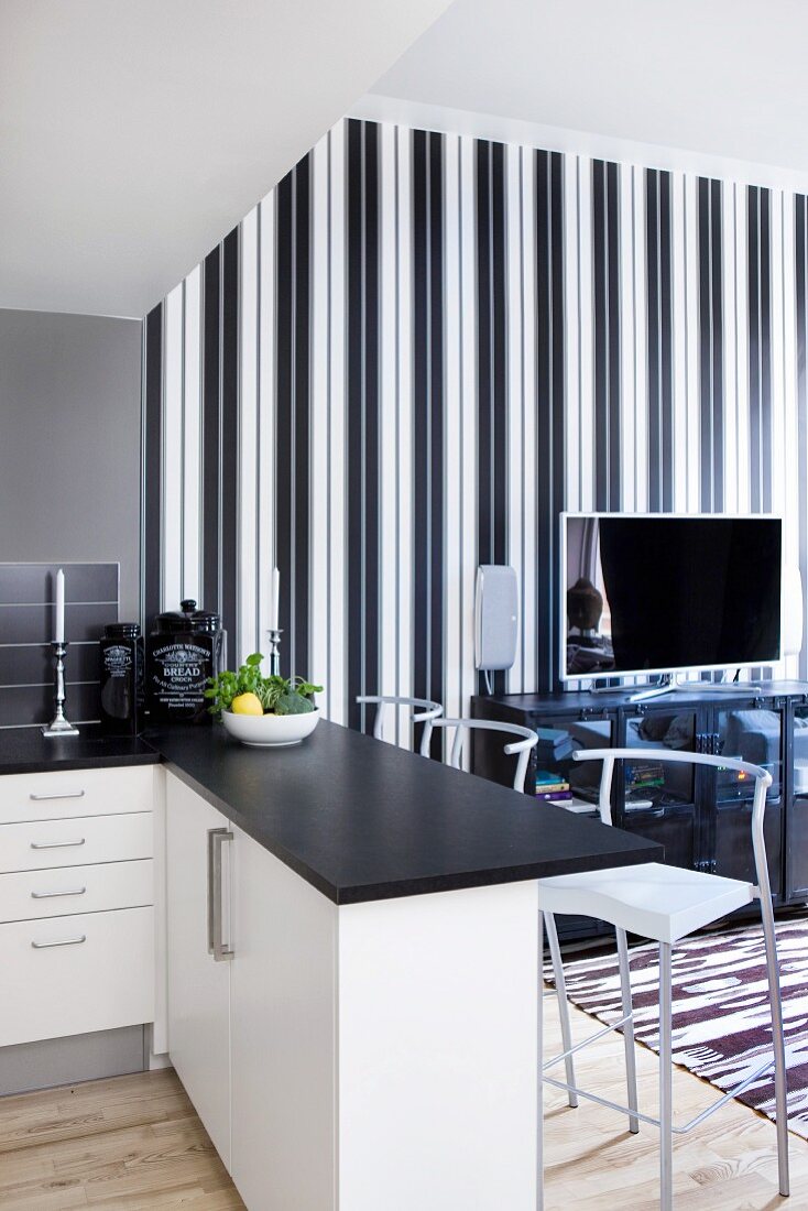 Offene Küche mit schwarzer Arbeitsplatte auf Esstheke und Barhocker, im Hintergrund Fernseher auf Sideboard vor eleganter, schwarzweisser Streifentapete