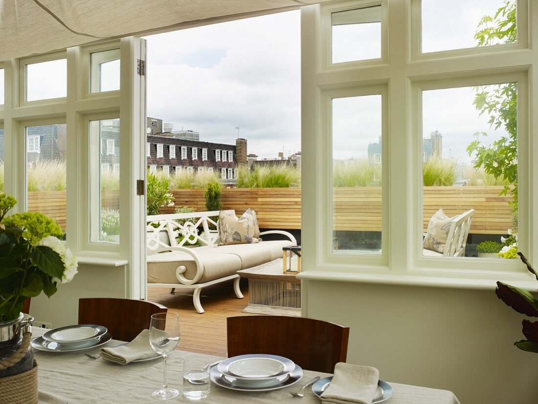 Gedeckter Tisch im Wintergarten mit Blick auf Dachterrasse und weiße nostalgische Sitzbank