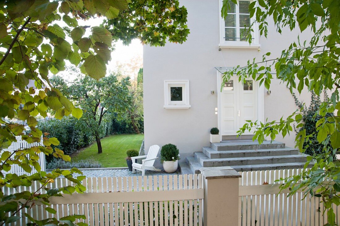 View over white picket fence of front door of house with stone steps and well-tended garden