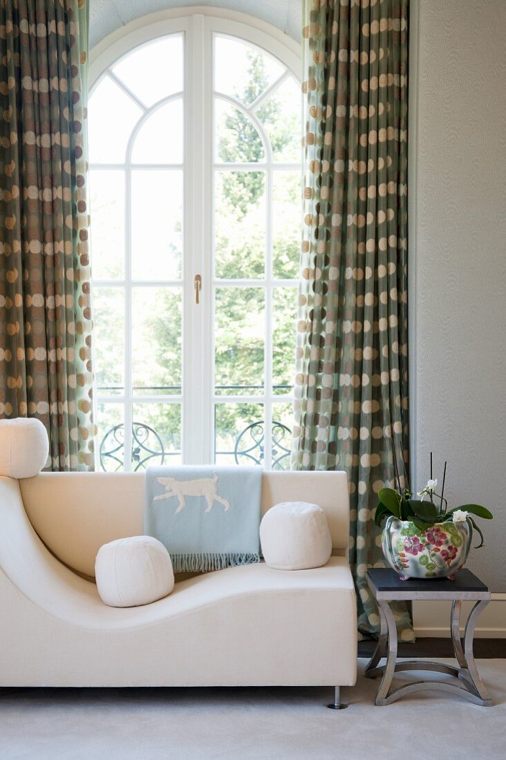 White, round cushions on modern sofa and potted orchid on side table in front of arched window
