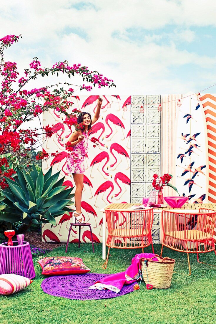 Flamingo-Prints, Outdoor-Möbel und Accessoires in Rot- und Lilatönen; Frau auf Hocker beim Aufhängen einer Lichterkette