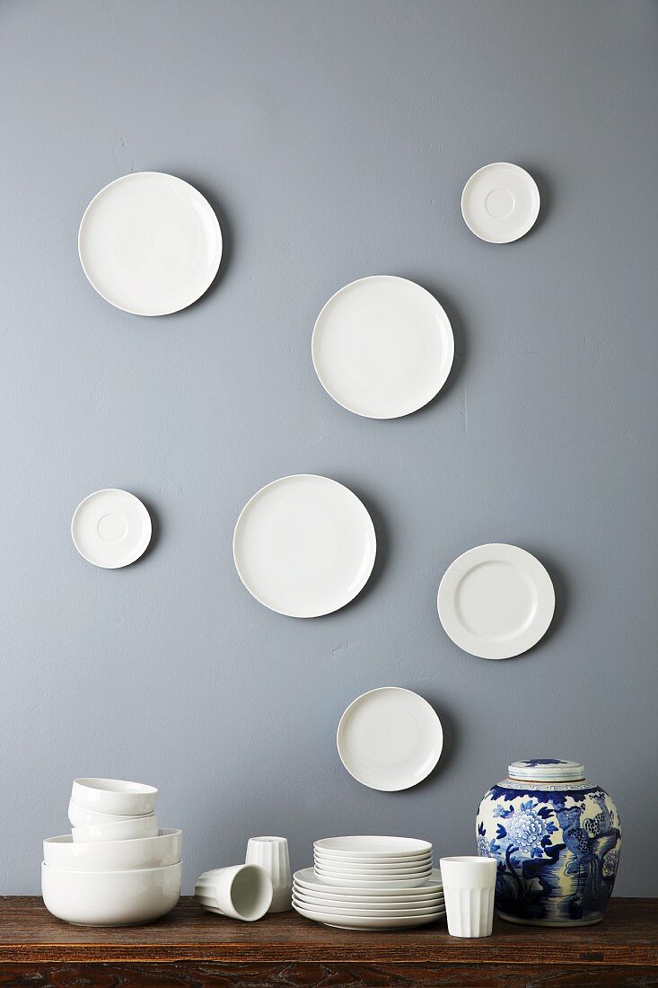 Gestapeltes, schlichtes Porzellan und gemusterte Deckelvase, darüber weiße Teller auf grauer Wand