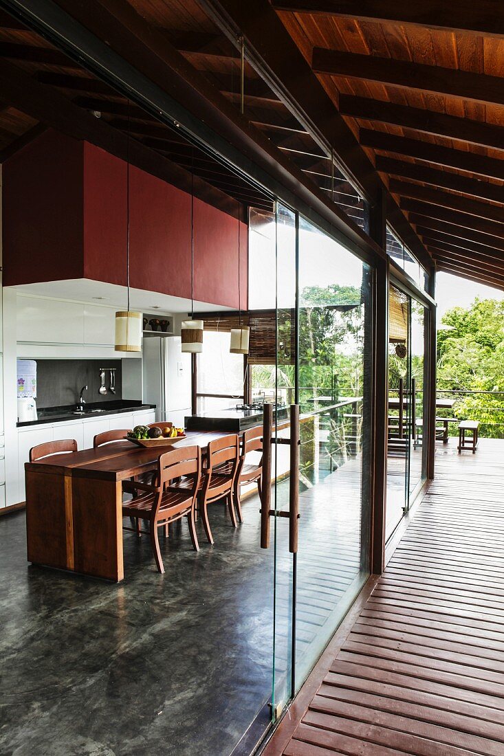 Frameless, sliding glass elements form transition between roofed wooden terrace and contemporary, open-plan kitchen with integrated dining area