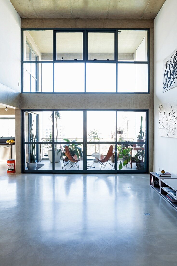 Blick von minimalistischem Wohnraum mit poliertem Betonboden auf überdachte Terrasse mit Butterfly Chairs hinter zweigeschossiger Glasfassade