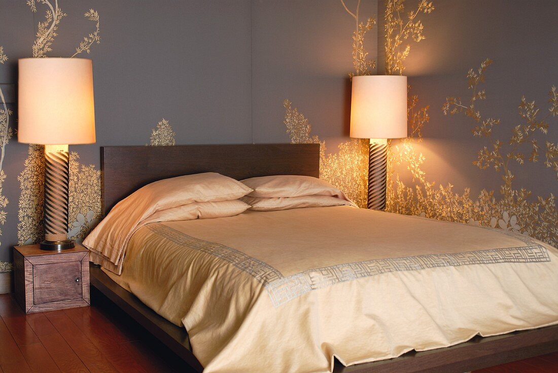 Elegant satin bed linen on double bed flanked by large bedside lamps on small wooden boxes against grey and gold, floral-patterned wallpaper