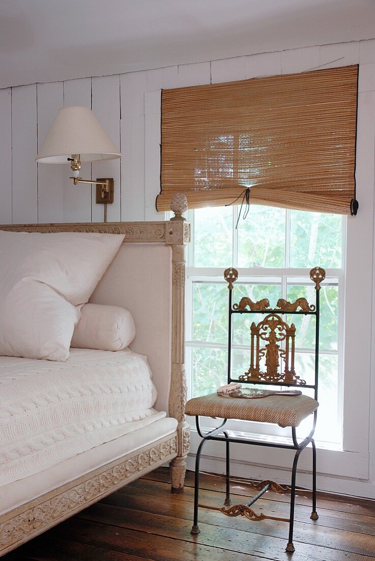 Filigraner Metallstuhl mit verzierungen neben antikem Bett mit hohem Kopfteil am Fenster mit halbgeschlossenem Rattan Rollo