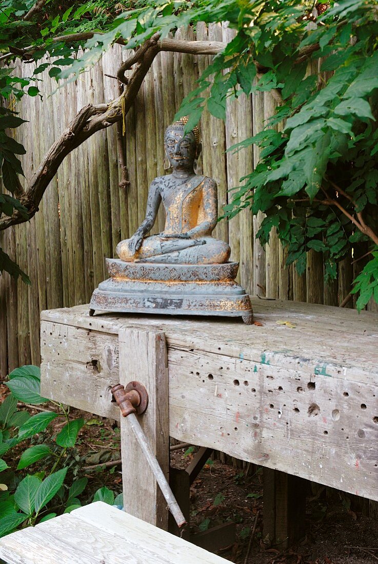 Buddhafigur aus Metall auf Vintage Werkbank in Freiem vor Holzzaun