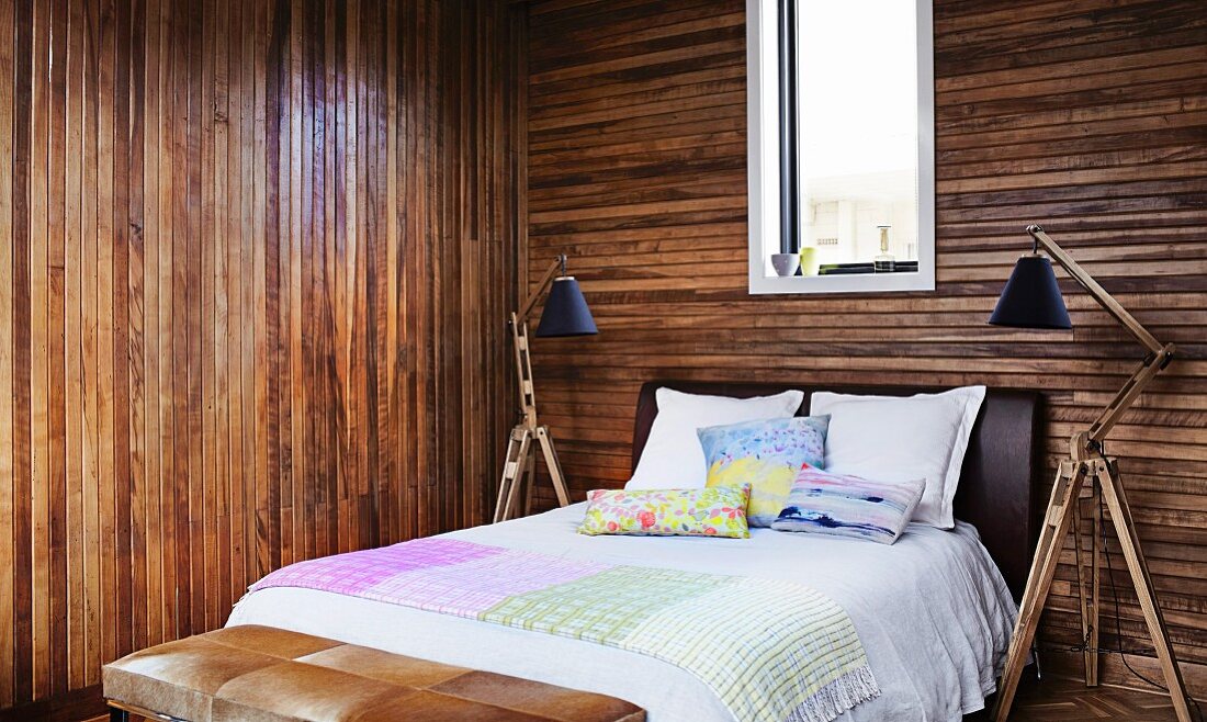Schlichtes Doppelbett zwischen Stehleuchten mit verstellbarem Holzgestell in Schlafzimmer mit dunklen Holzwänden