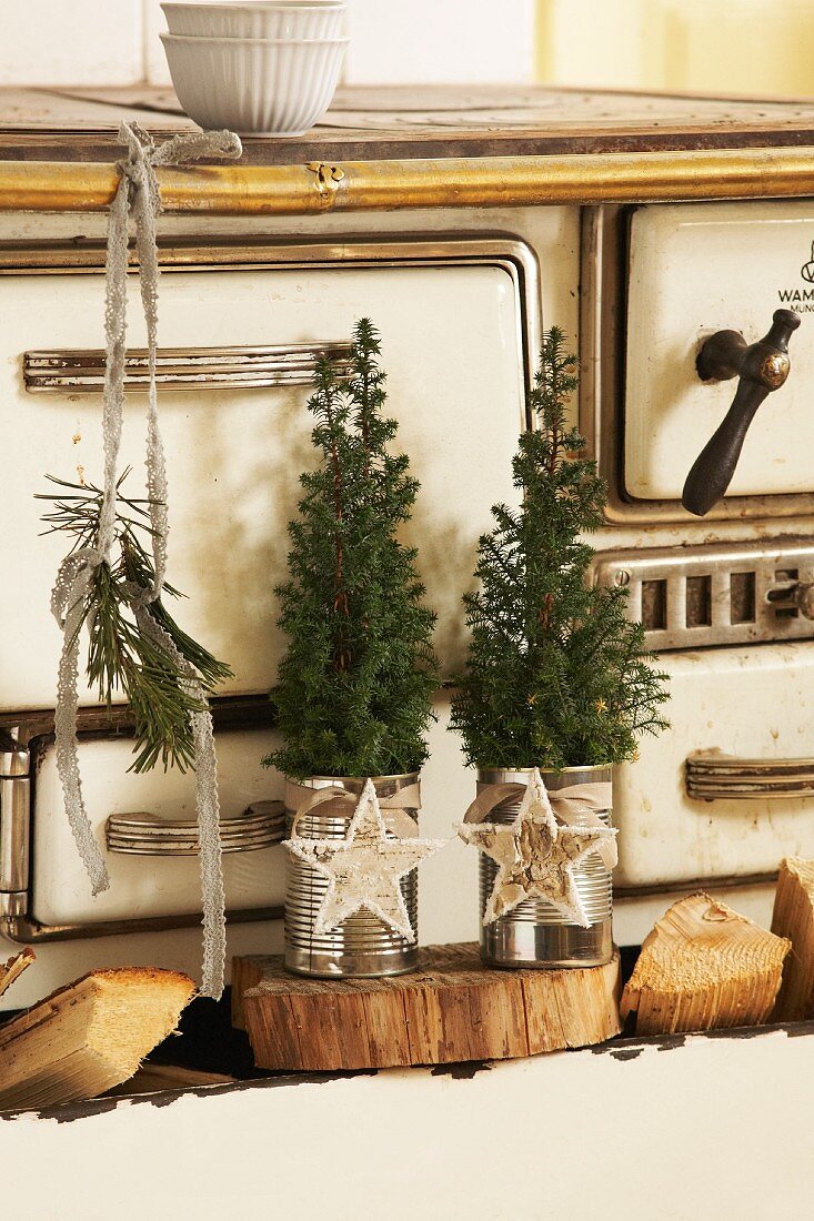 Small fir trees in tin cans decorated with Christmas baubles on wooden board in front of vintage kitchen cooker