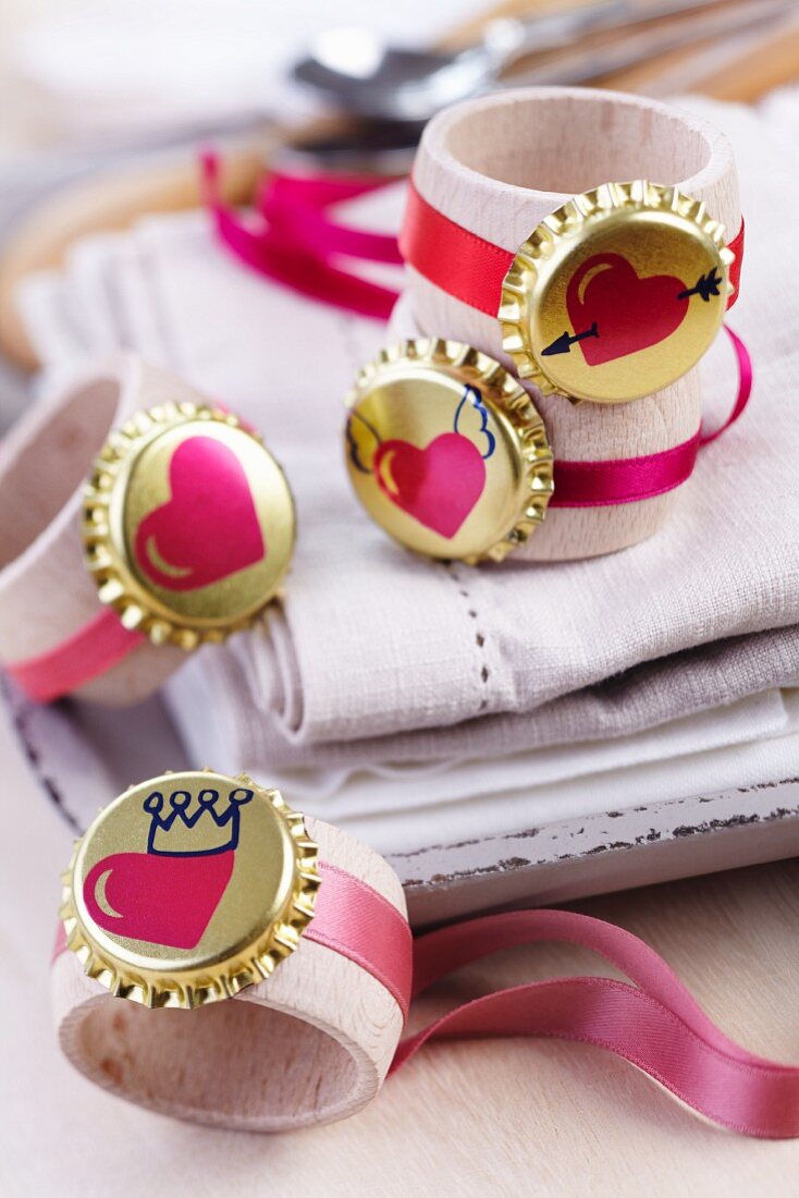 Goldfarbene Kronkorken mit Herzmotiven und rosafarbenen Geschenkbändern an Holzserviettenringen