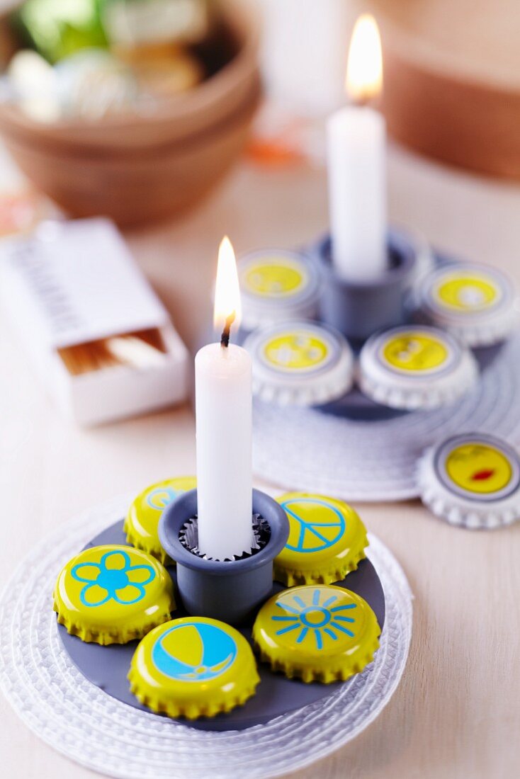 Simple candle stick and bottle caps painted yellow with various motifs arranged on white coaster