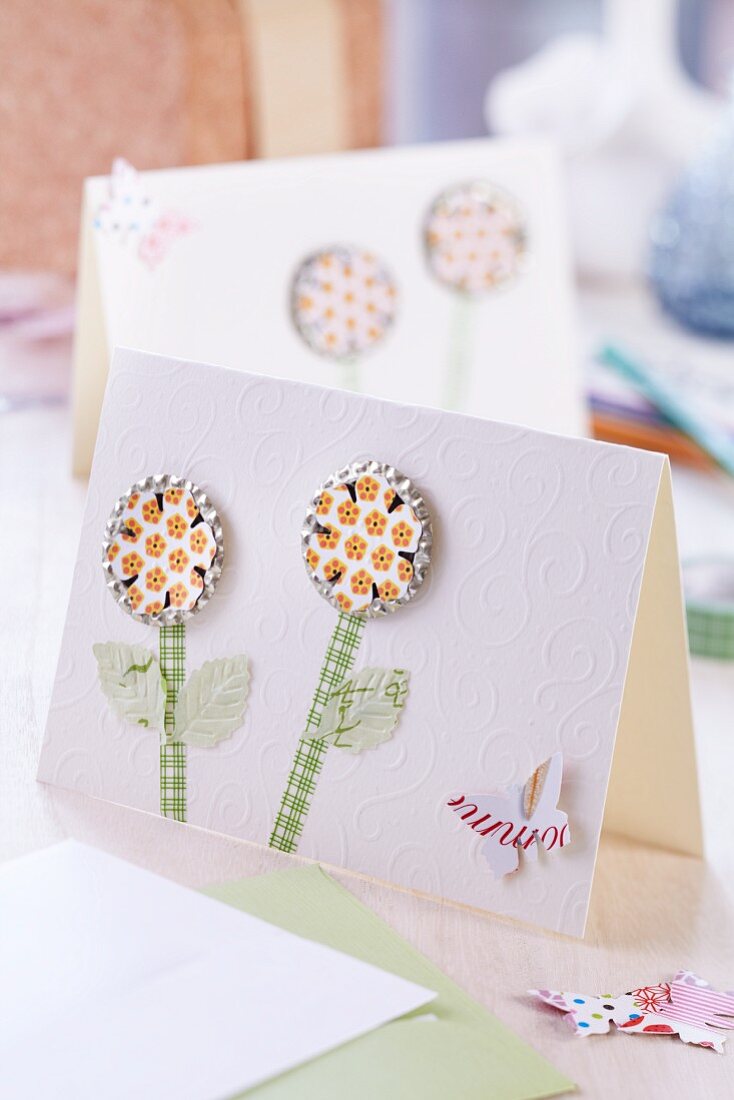 Flachgedrückte beklebte Kronkorken als Blütenmotiv mit Papierstengel und Tiermotiv auf edler weißer Glückwunschkarte