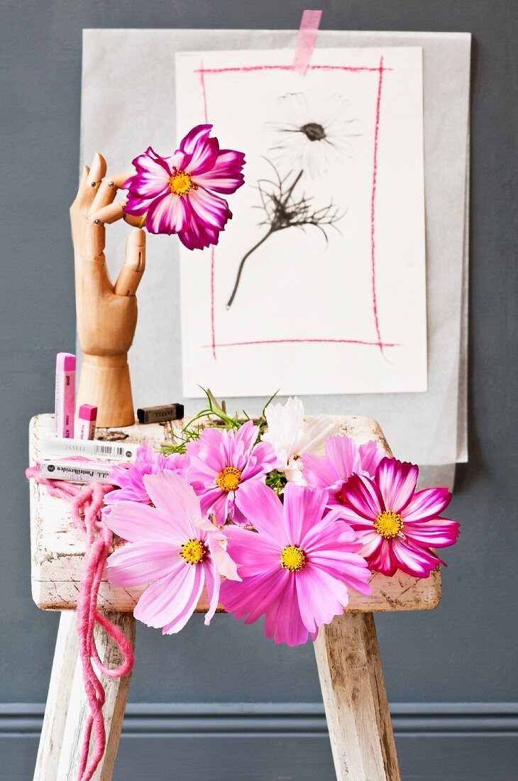 Small still-life arrangement of various cosmos flowers, pastel chalks and wooden jointed hand with floral drawing on grey wall in background