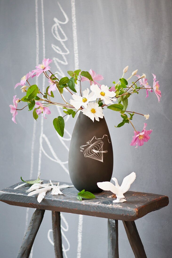 Dunkelgraue Retro Vase mit Cosmea-Strauss auf altem Holzhocker vor grauer Wand