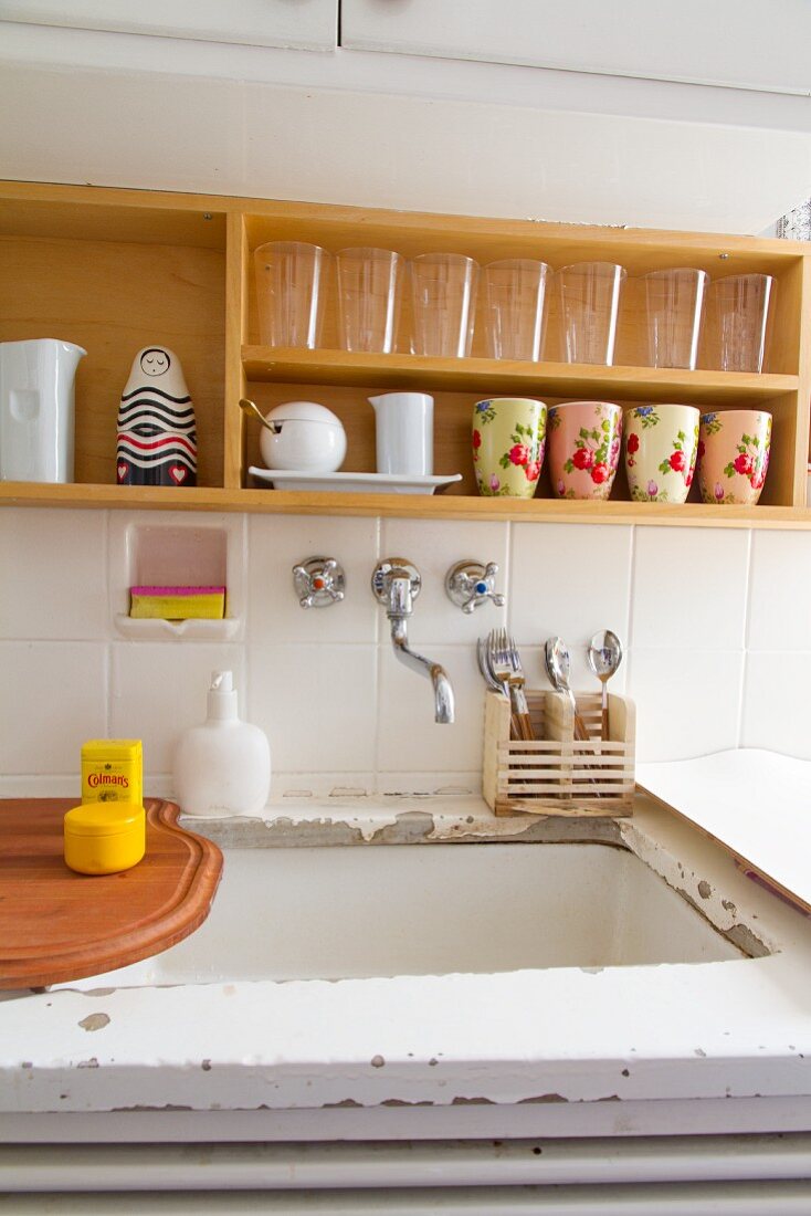 Glasses and painted crockery on wall-mounted wooden shelves above sink and white worksurface with peeling paint