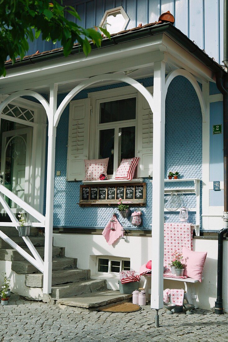 Romantic entrance to renovated country house with pale blue clapboard facade and red and white cushions on white bench