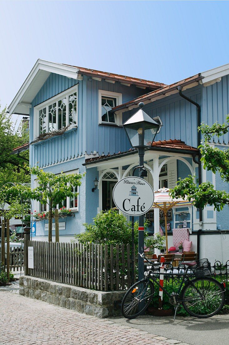 Blick auf restauriertes Landhaus- Café mit hellblauer Holzfassade, idyllischem Vorgarten und lauschigem Terrassenplatz