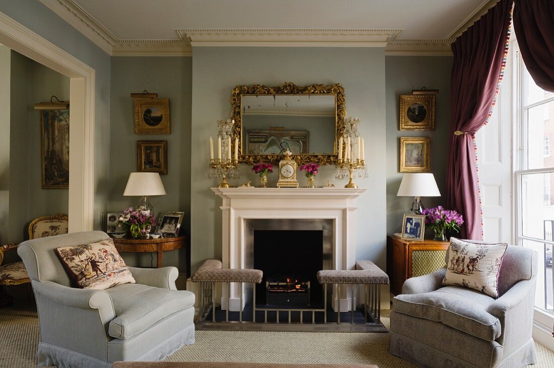 Wide armchairs in front of English fireplace with ornaments on mantelpiece and fender seat