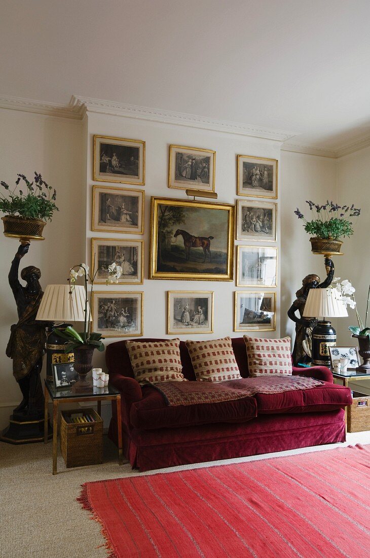 Gilt-framed, historical paintings above red velvet sofa flanked by side tables and statues holding plants on trays
